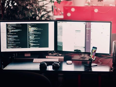 Schreibtisch mit zwei Monitoren, links Programmcode, davor Tastatur, Kopfhörer und Smartphone im Ständer