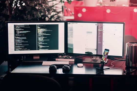 Schreibtisch mit zwei Monitoren, links Programmcode, davor Tastatur, Kopfhörer und Smartphone im Ständer