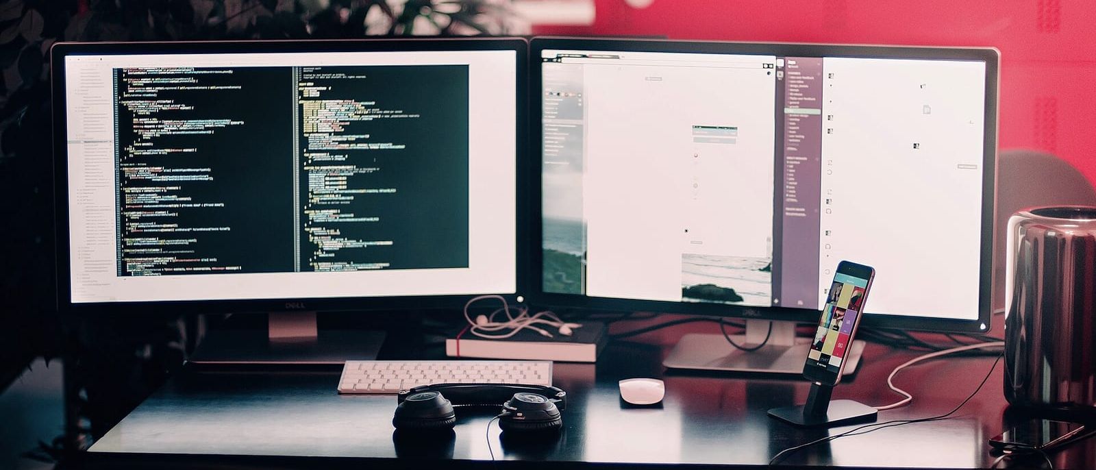 Schreibtisch mit zwei Monitoren, links Programmcode, davor Tastatur, Kopfhörer und Smartphone im Ständer