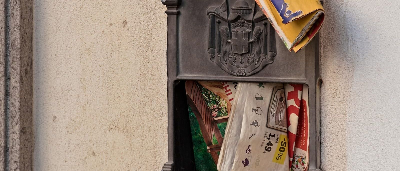 Überquellender metallener Briefkasten an einer Hauswand, Prospekte ragen heraus