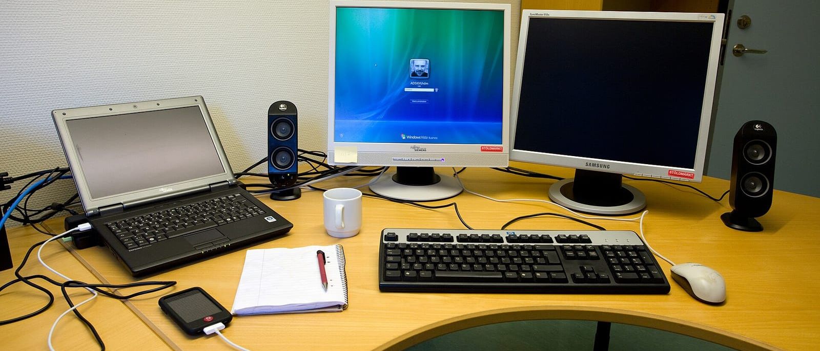 Büroschreibtisch mit Laptop, zwei Monitoren, Tastatur, Notizblock und Kaffeetasse