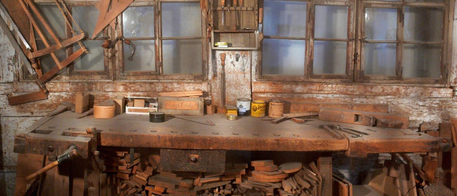 Alte Schreinerwerkstatt mit Hobelbank, Holzresten und Werkzeug vor Sprossenfenstern